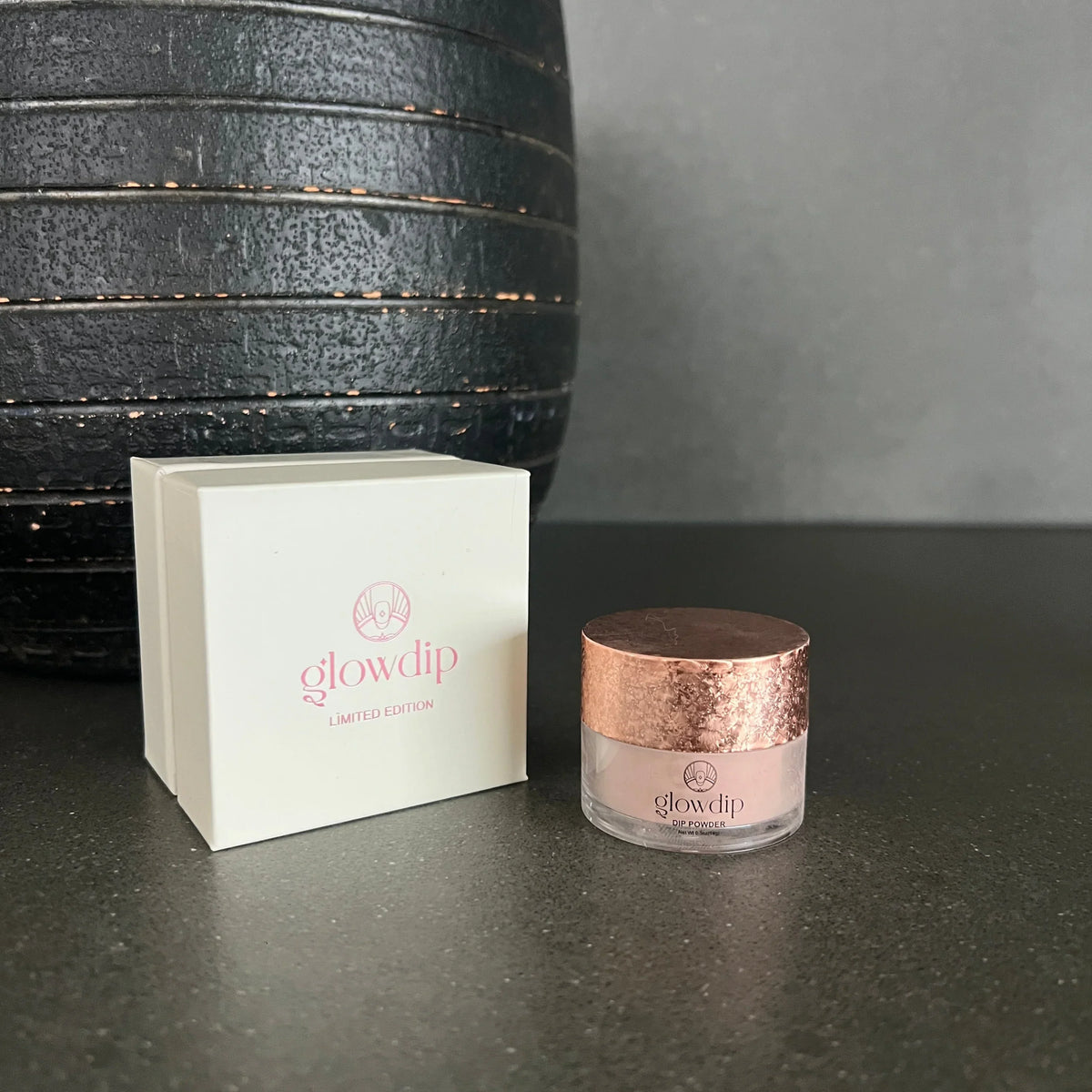 Een rose gouden textuurde deksel bedekt een glazen pot met lichtroze glowdip poeder naast een witte limited edition doos