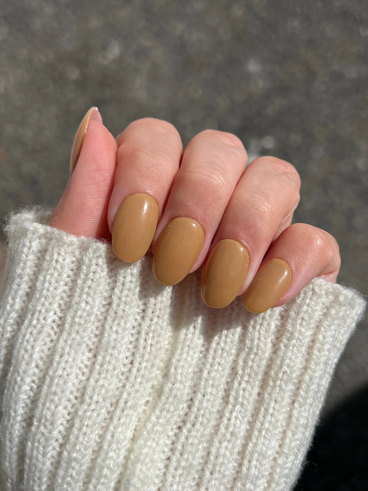 Een hand met netjes gemanicuurde nagels in matte karamelkleurige golden drift dipping powder en een zachte witte ribgebreide trui